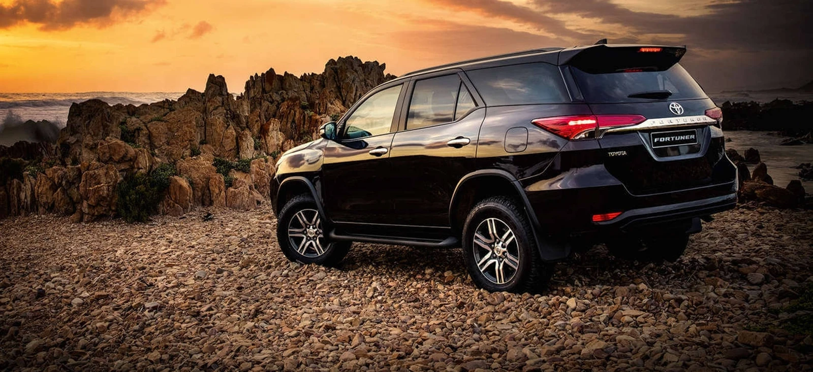 Toyota Fortuner beside a rocky coast at sunset.
