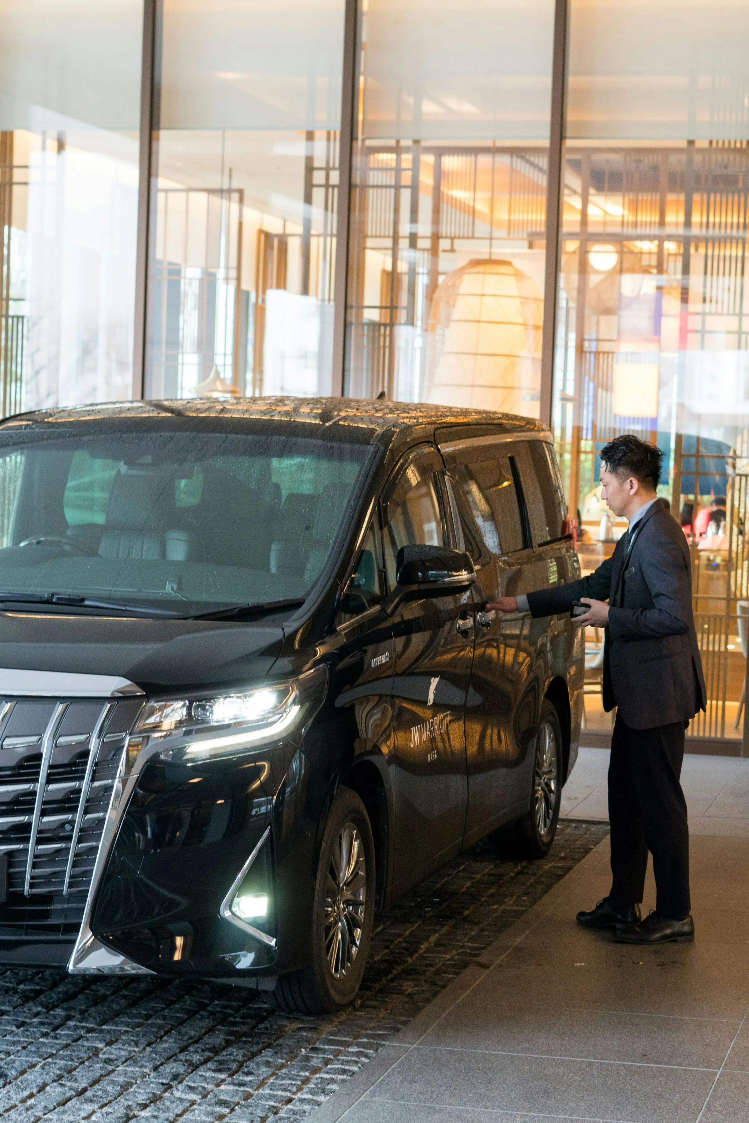 A chauffeur in a dark suit opening the rear door of a black premium people-mover outside a warmly lit hotel lobby at dusk.