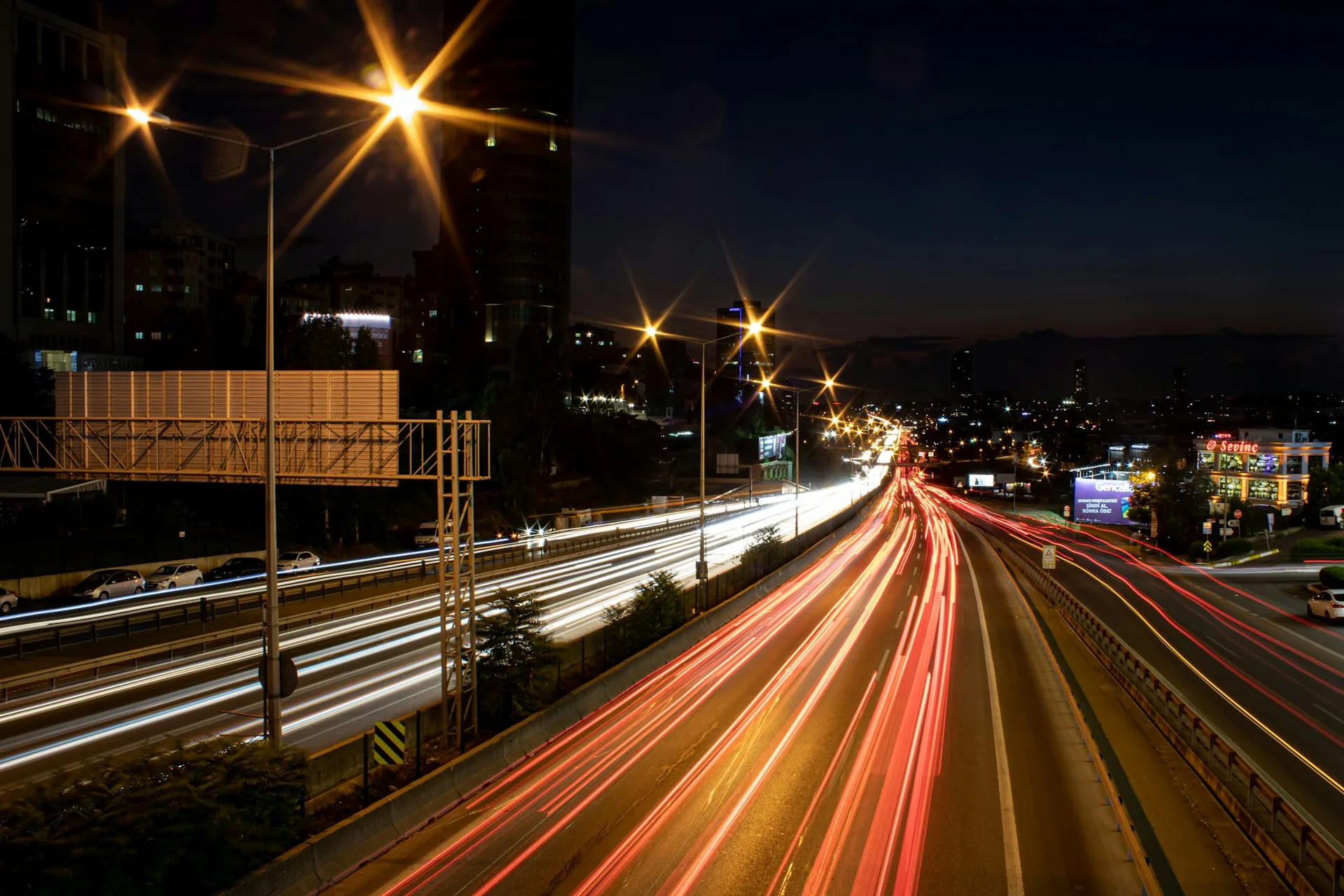A lit highway curving away from a city at night, headlights drawn into long trails.