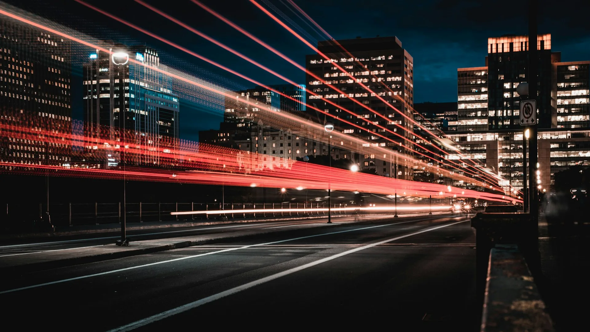 Light trails from traffic streaking past office towers on a city expressway at night.